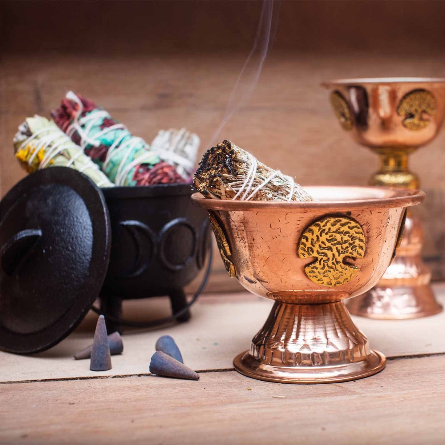 Copper incense burner with smoldering incense sticks on a wooden surface, surrounded by other incense and tools.